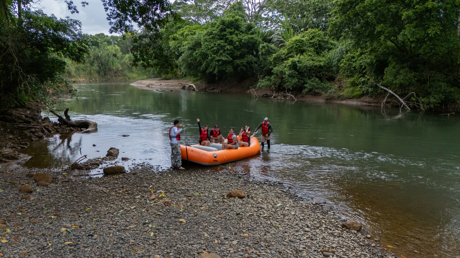 Vista aérea baja del momento de embarque — raft naranja amarrado en la orilla rocosa del Río Peñas Blancas con grupo subiendo